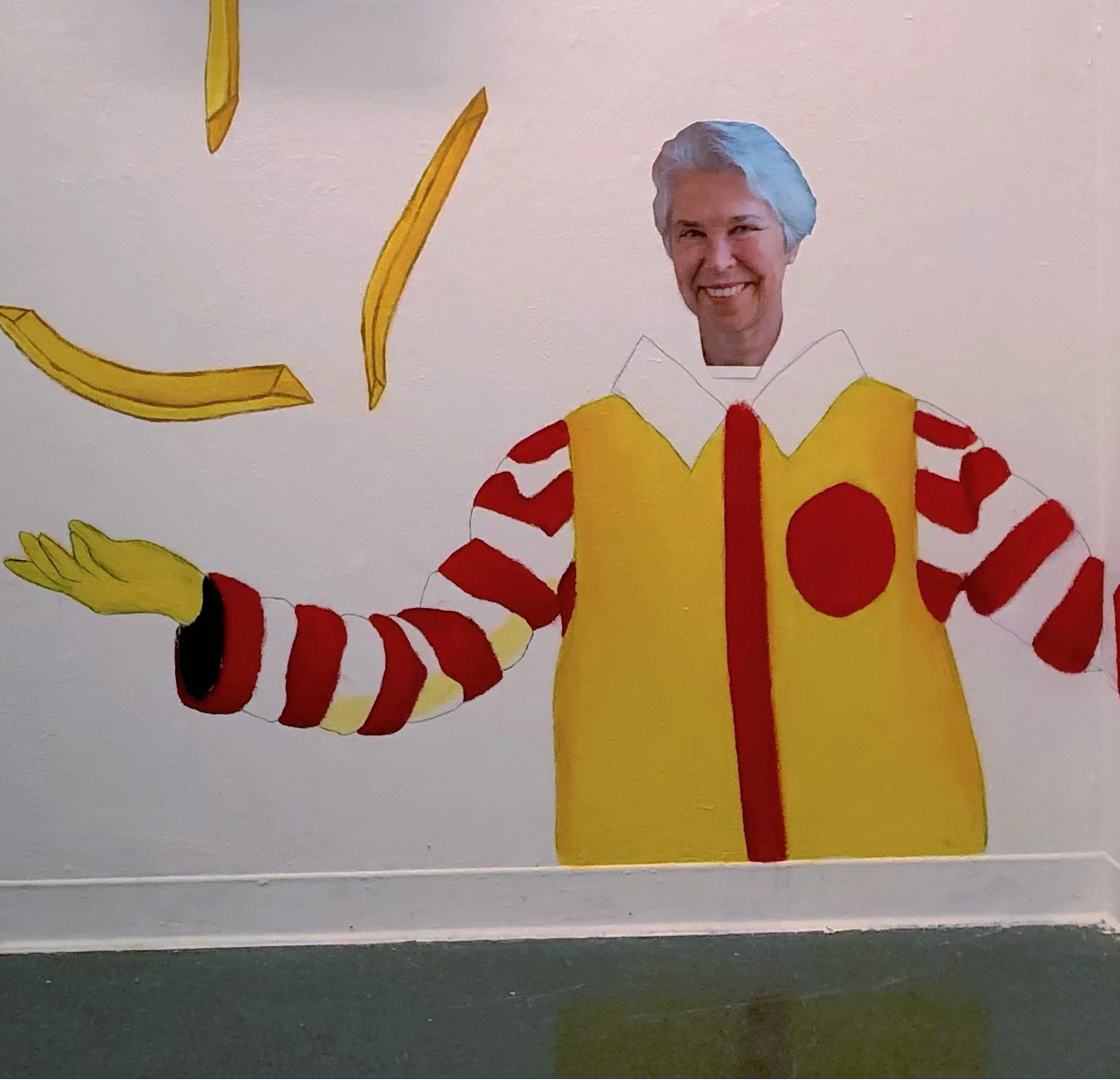 A wall-mounted art installation in a gallery space featuring a stylized golden arches-themed mural with french fries, a red and yellow logo shape, two black wall-mounted screens, and a cutout figure of Ronald McDonald on a white wall.